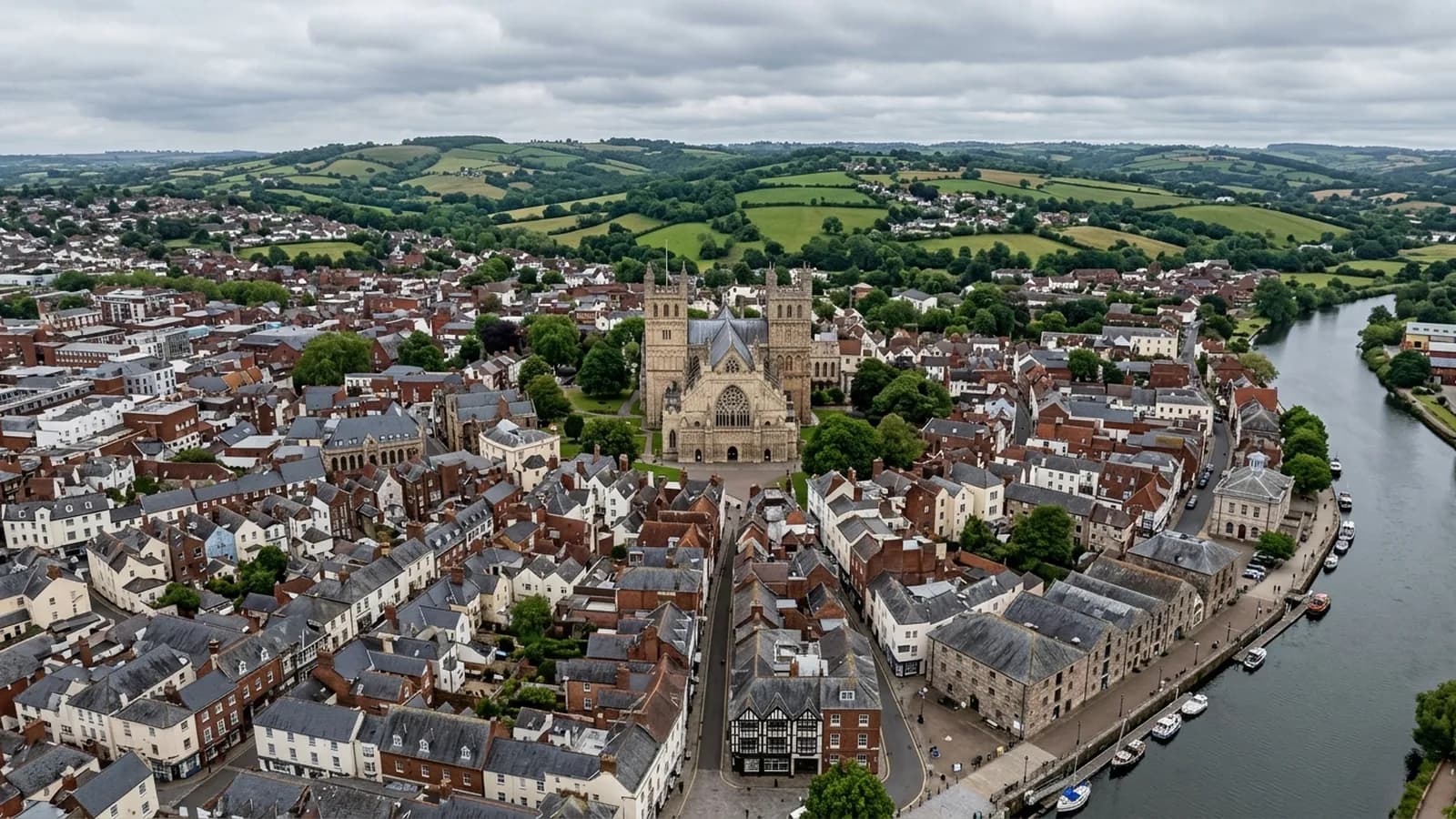 Aerial view over a Devon cathedral city on a grey afternoon, dense slate and tile rooftops packed close together with a tower rising above them and a river along one edge